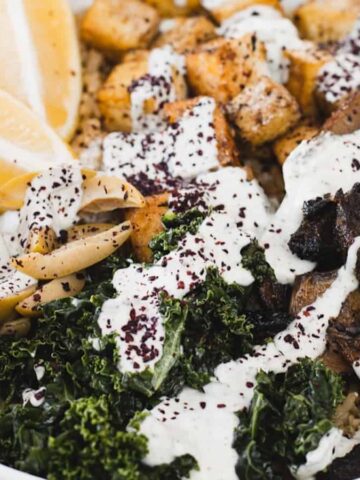 A bowl of mixed ingredients including lemon wedges, roasted tofu, mushrooms, kale, and rice, topped with a creamy white sauce and sprinkled with seasonings.