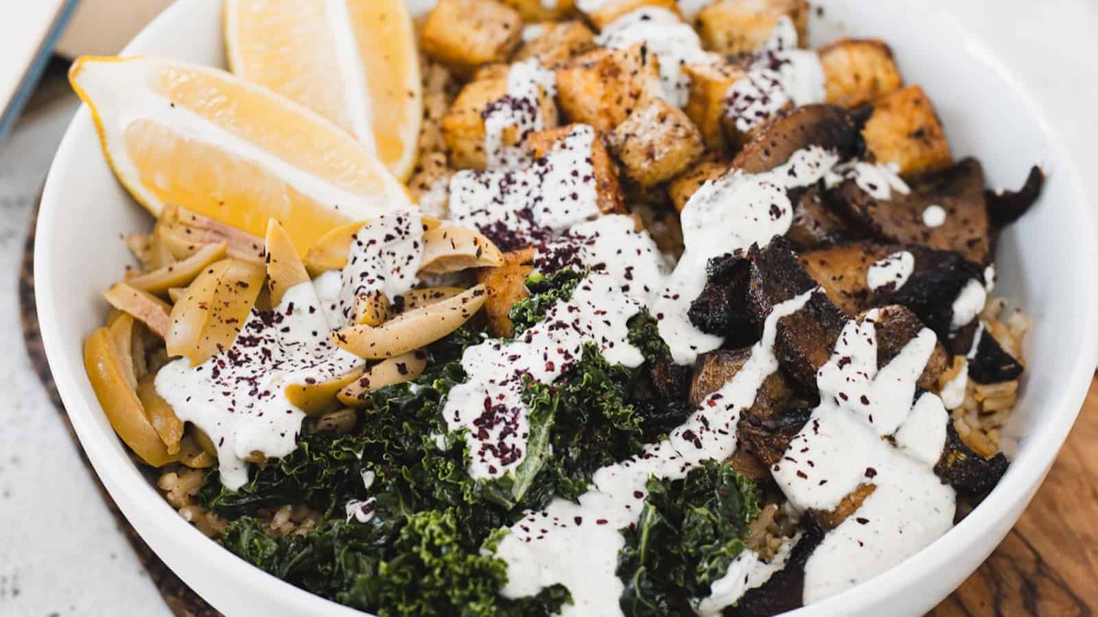A bowl of mixed ingredients including lemon wedges, roasted tofu, mushrooms, kale, and rice, topped with a creamy white sauce and sprinkled with seasonings.