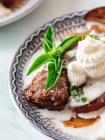 A dessert plate with a slice of cake topped with whipped cream and a sprig of fresh basil. The cake is surrounded by a sweet sauce and served in a patterned dish with a spoon beside it. Another plate is partially visible in the background.