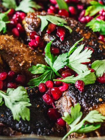 A dish featuring braised meat garnished with fresh parsley leaves and scattered pomegranate seeds. The meat appears to have a caramelized or charred surface, and the dish is presented in a rustic pan.