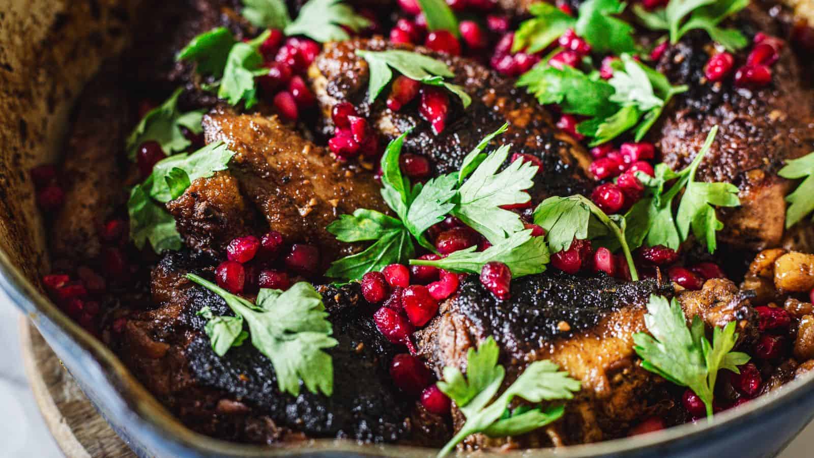 A dish featuring braised meat garnished with fresh parsley leaves and scattered pomegranate seeds. The meat appears to have a caramelized or charred surface, and the dish is presented in a rustic pan.
