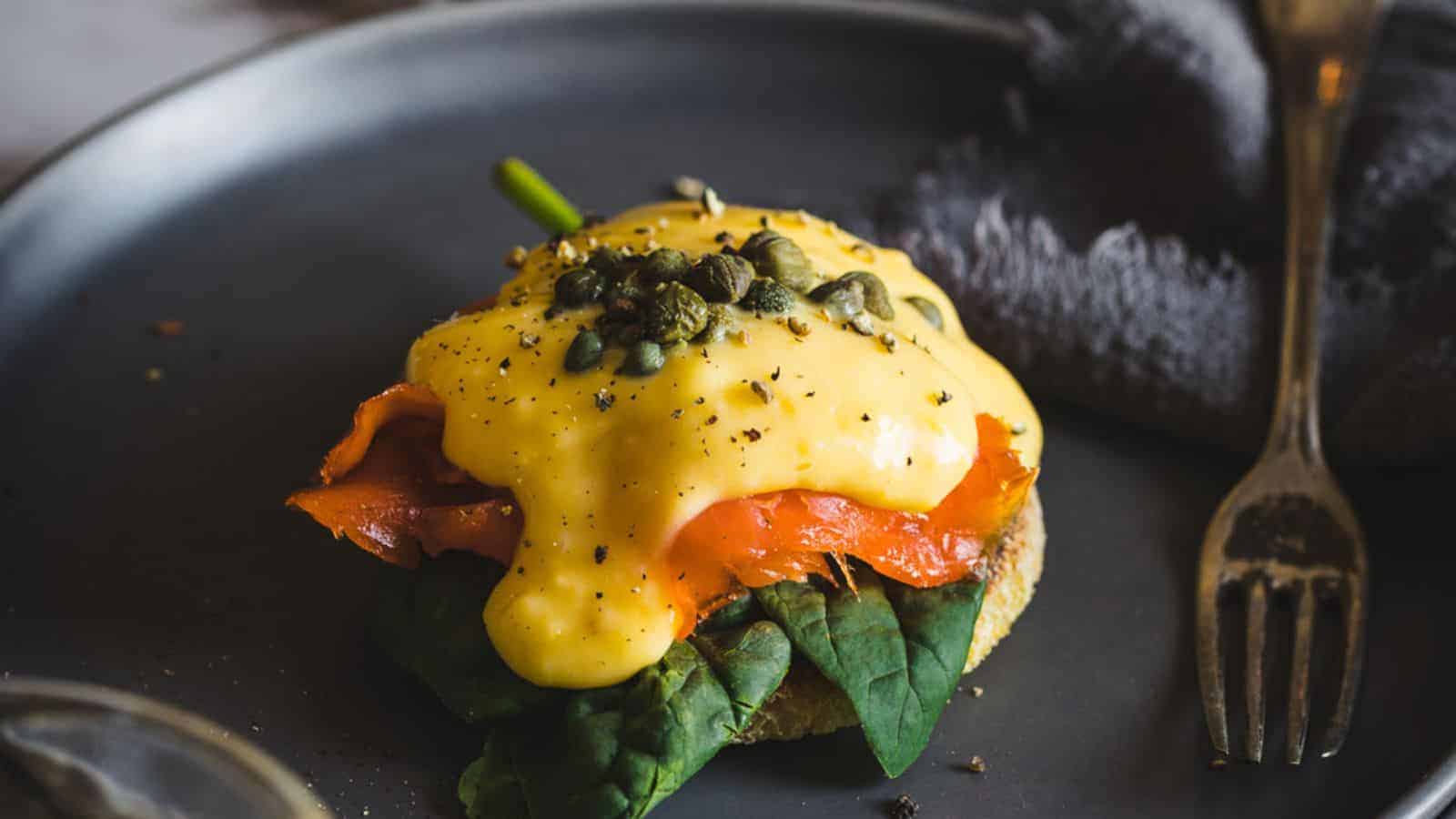 A person holding an eggs benedict with salmon and capers on a plate.