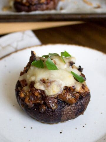 A stuffed mushroom topped with melted cheese and garnished with green herbs is centered on a speckled white plate. The plate rests on a wooden surface, and a tray with more stuffed mushrooms is in the background.
