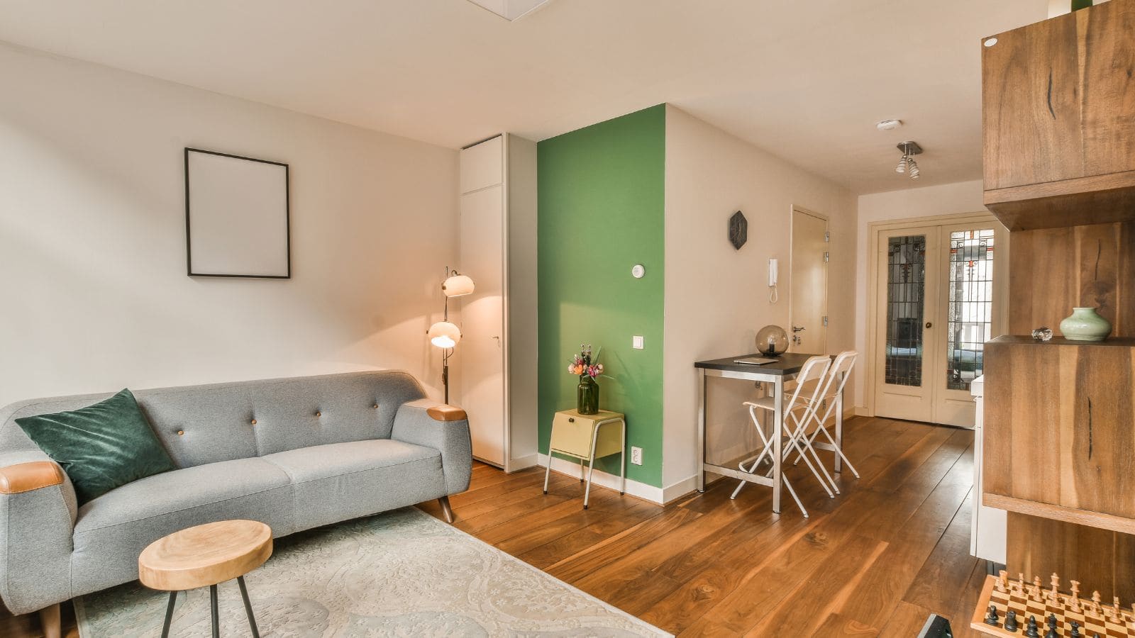 A cozy living room features a gray sofa with a green pillow, a wooden side table, and a standing lamp. A small dining area with a white table and chairs sits against a green accent wall. A shelf and decorative items are seen on the right.