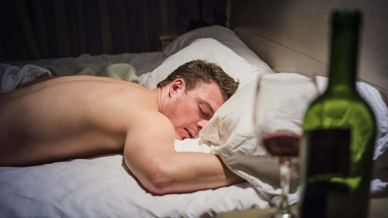A shirtless man is asleep in bed, lying face down on a pillow. In the foreground, there is an empty wine glass and a wine bottle on a nearby surface. The room is dimly lit.