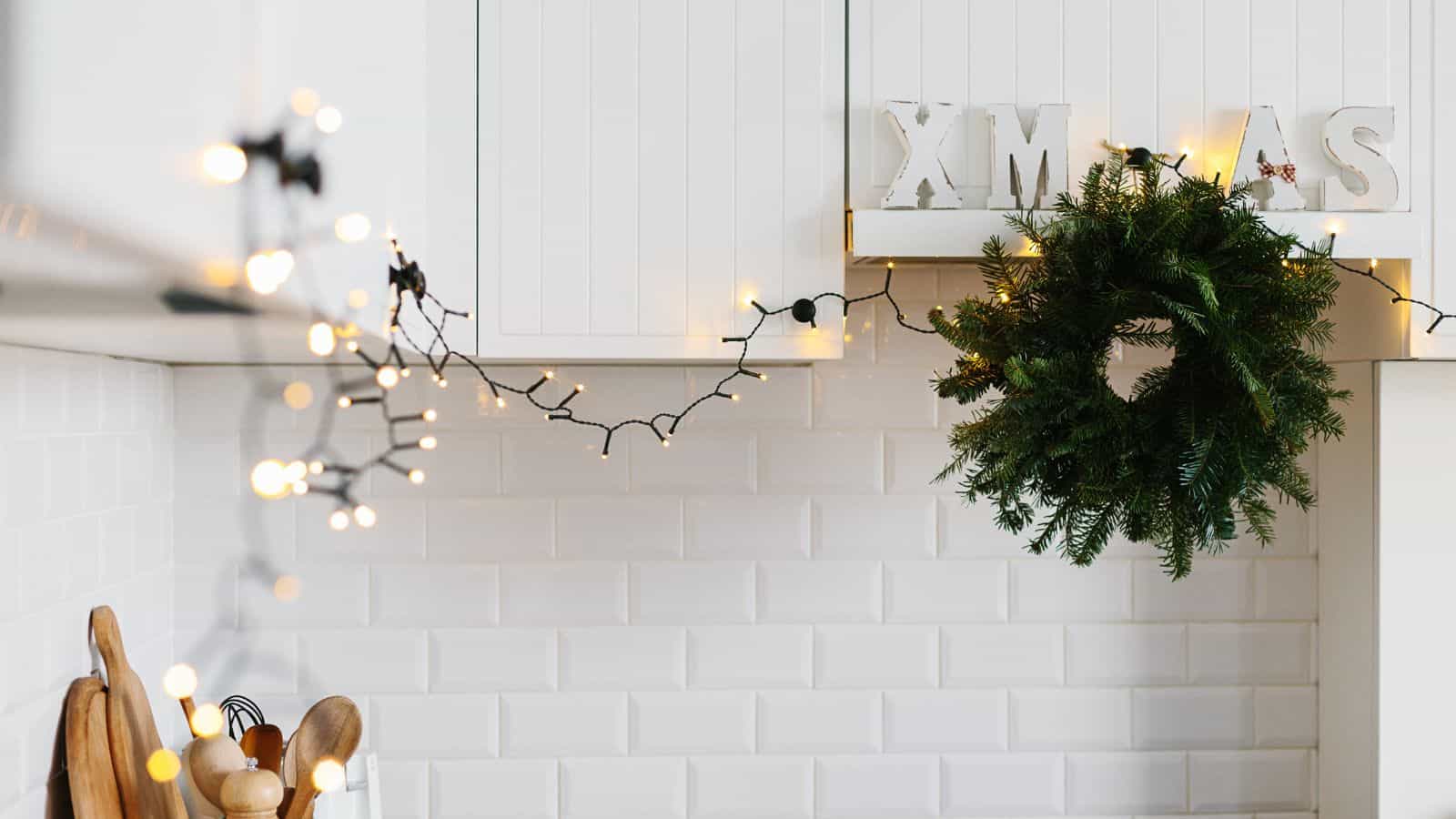 A kitchen with white tile walls decorated for Christmas. A green wreath hangs by a cabinet, and a string of warm white lights drapes across. Wooden utensils sit in a holder on the countertop, and block letters spell "XMAS" above.