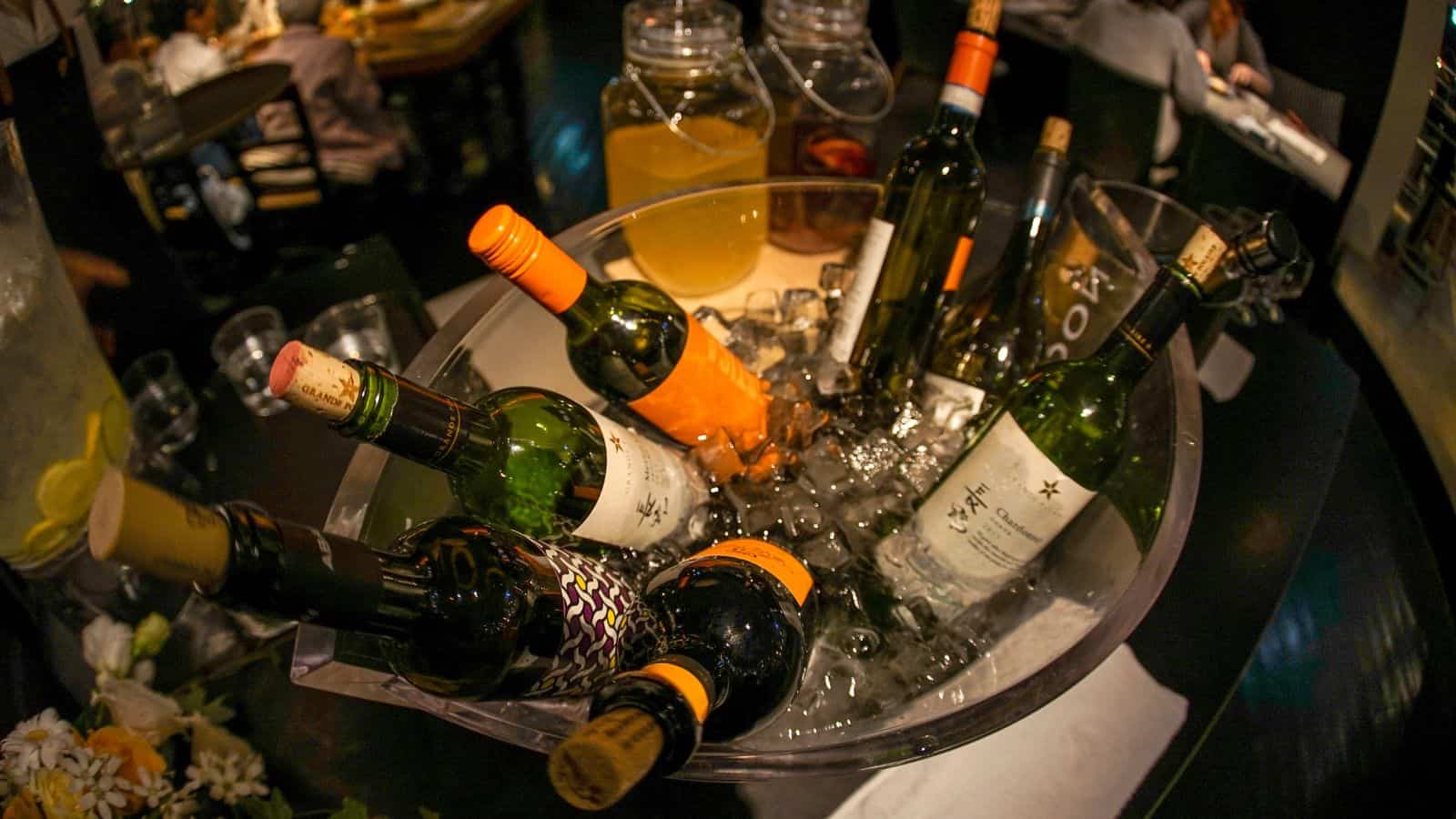 A large bowl filled with ice holds several bottles of wine. Two jars of juice and some glasses are in the background on a dark table.