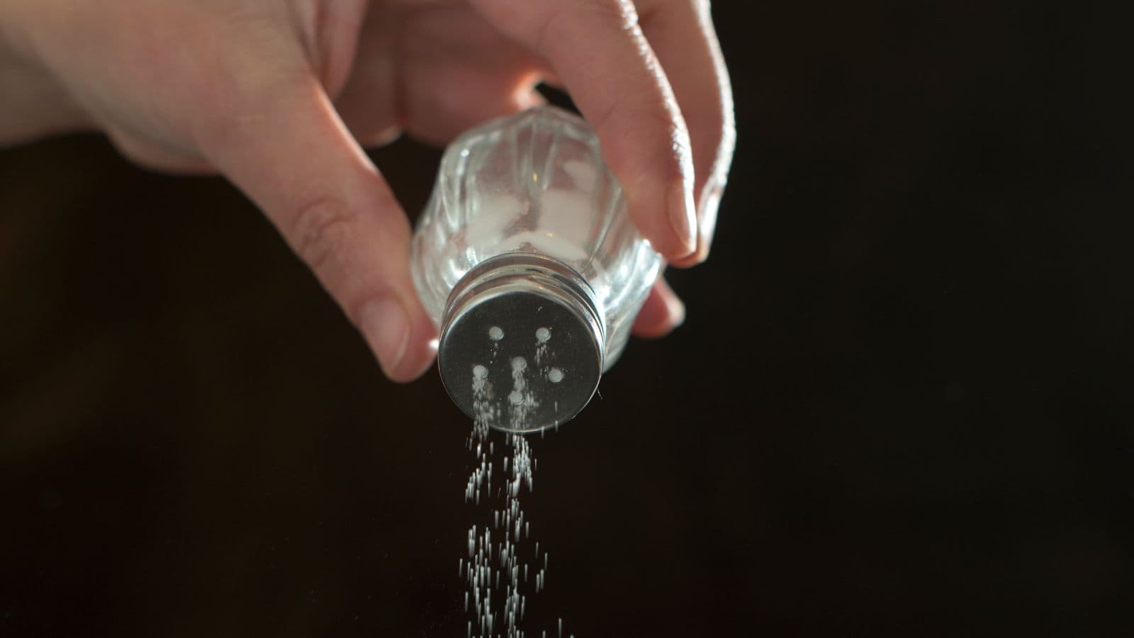 A hand is holding a salt shaker, sprinkling salt downwards against a dark background. The image captures the movement of the salt grains as they fall.