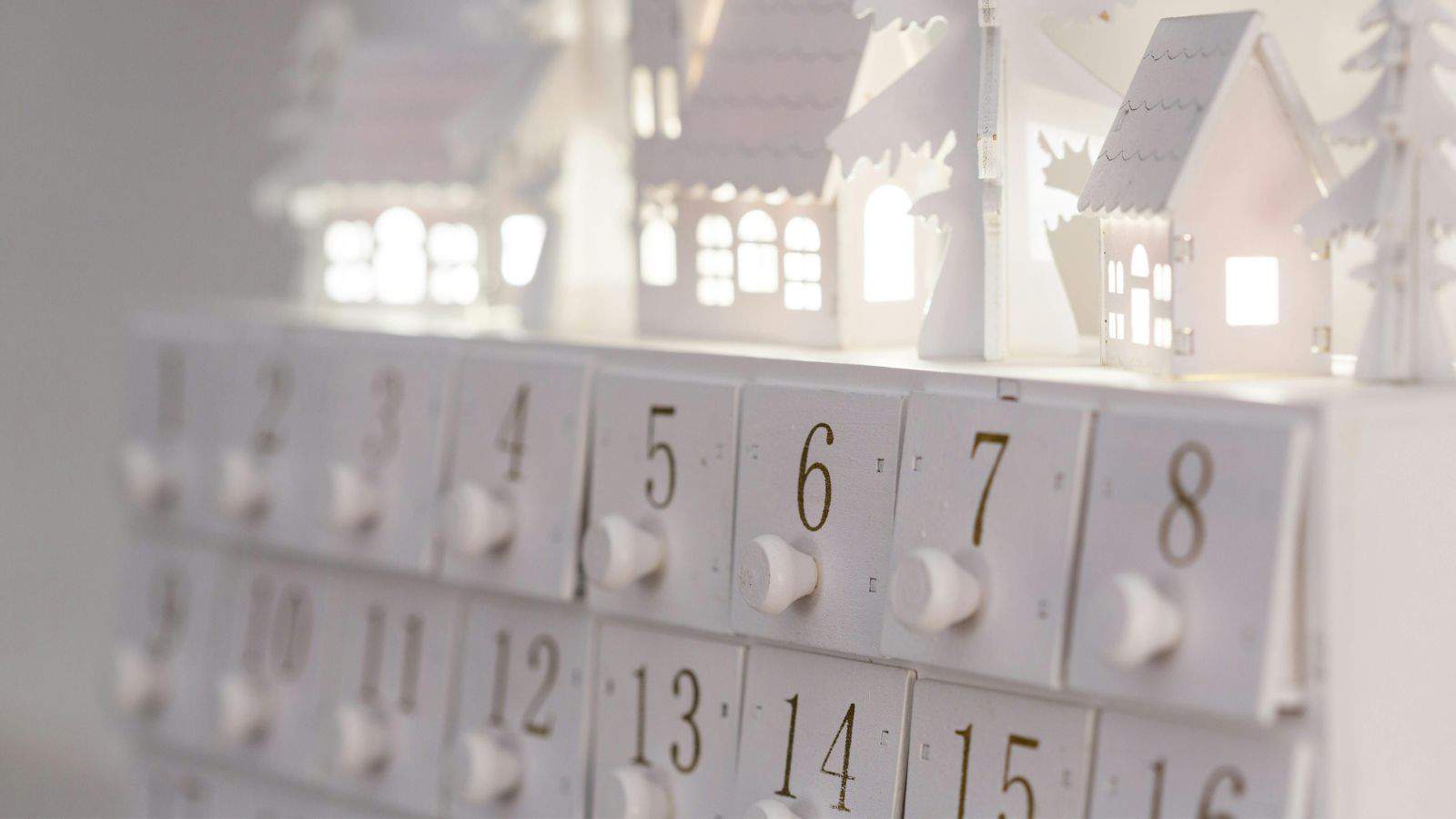 A white advent calendar featuring small numbered drawers from 1 to 15. Above the drawers are decorative silhouettes of houses and trees. The scene is softly illuminated from behind, casting a gentle glow on the white structures.