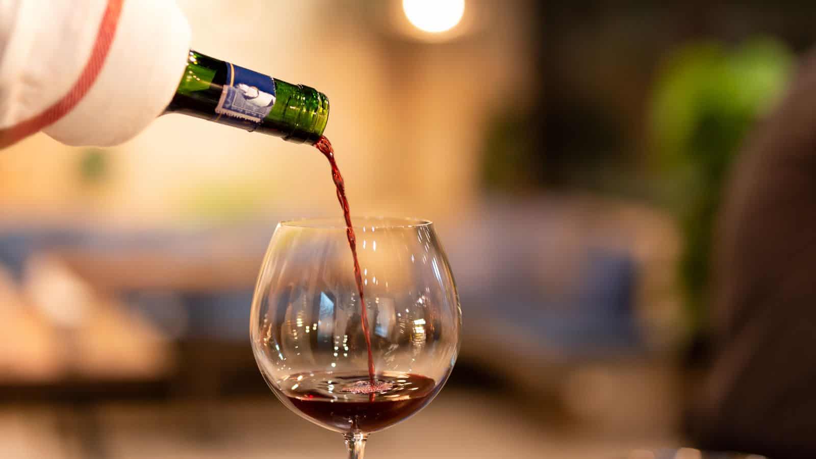 A close-up of red wine being poured from a green bottle into a clear wine glass. The background is softly blurred, highlighting the focus on the wine.