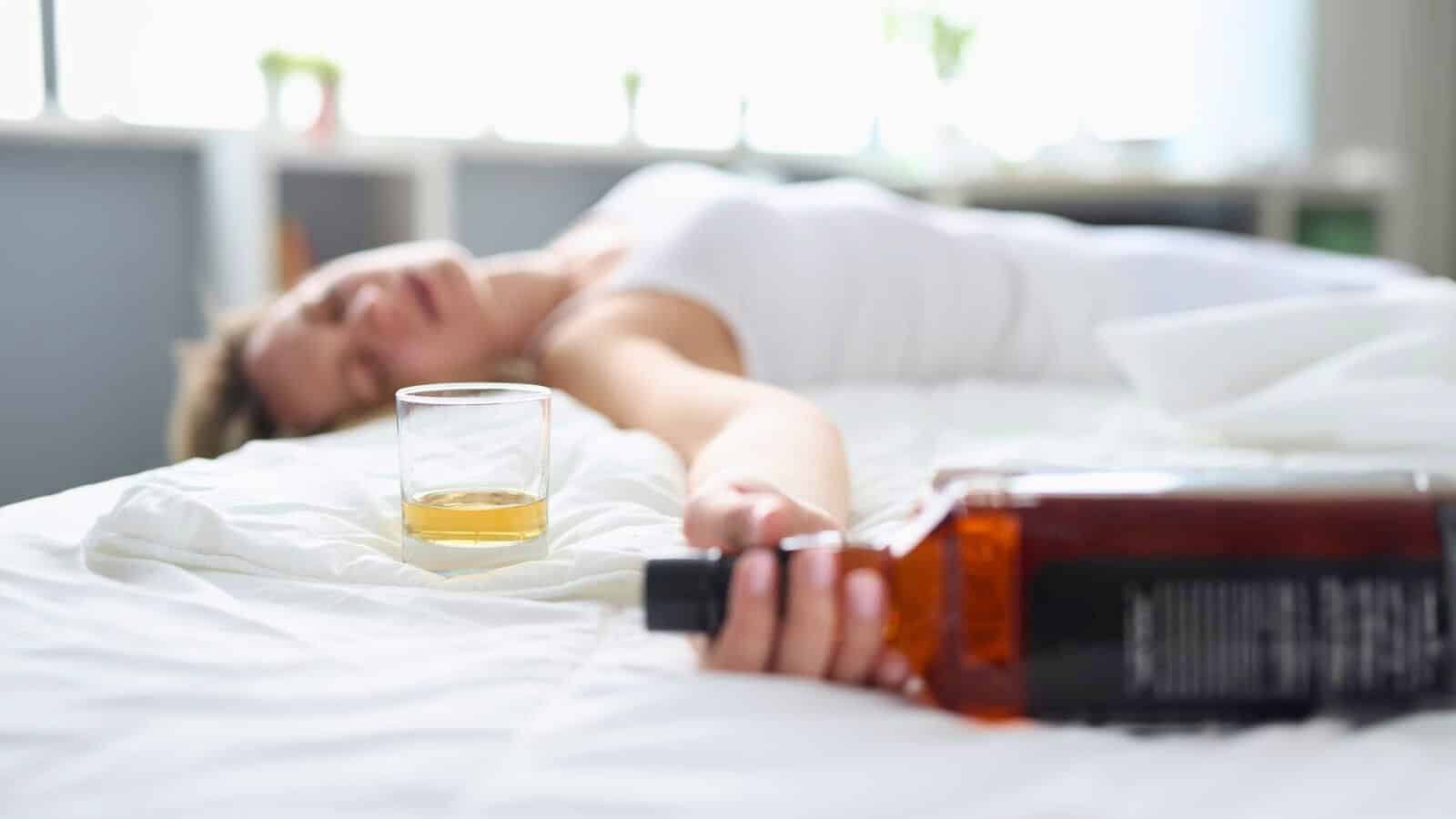 A person lies on a bed with a glass of whiskey and a bottle nearby. The person appears to be asleep or unconscious, with an outstretched arm reaching towards the bottle. The setting suggests a scene of relaxation or excessive drinking.