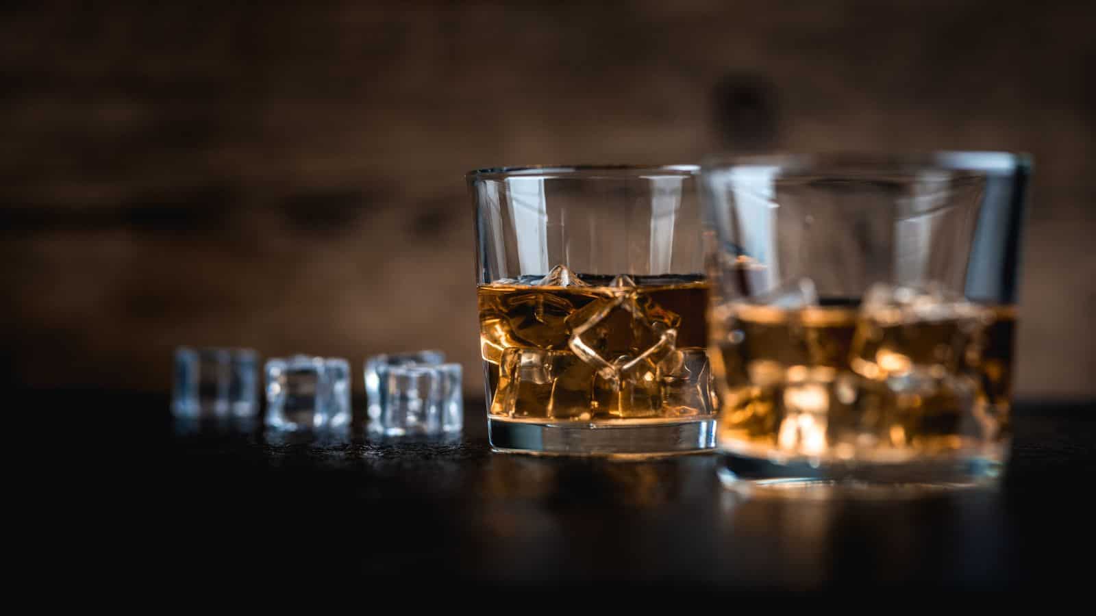 Two glasses filled with amber liquid and ice are in focus in the foreground. In the background, several ice cubes are lined up on a dark surface. The background is blurred and has a warm, rustic appearance.