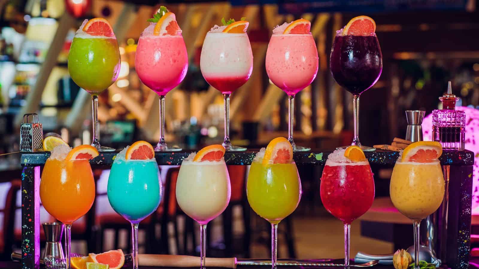 A colorful array of twelve cocktails is displayed, each garnished with a slice of orange. The drinks vary in bright colors, including green, pink, red, blue, yellow, and white, set on a bar counter with blurred bar stools and lights in the background.