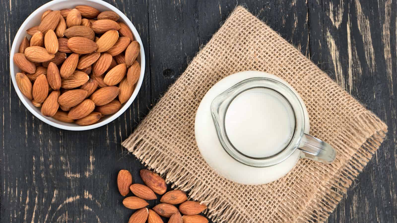 A bowl of almonds is placed on a dark wooden surface next to a small glass pitcher filled with milk. Both items sit on a square piece of burlap fabric, with a few almonds scattered on the wood.