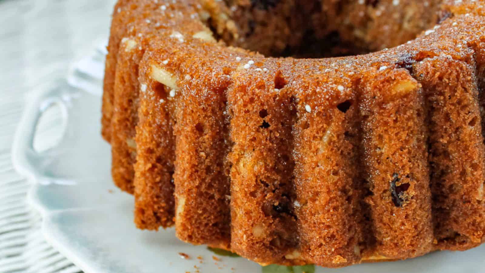 Close-up of a bundt cake on a white plate. The cake is golden brown with visible nuts and raisins, featuring a ridged texture and a moist appearance.