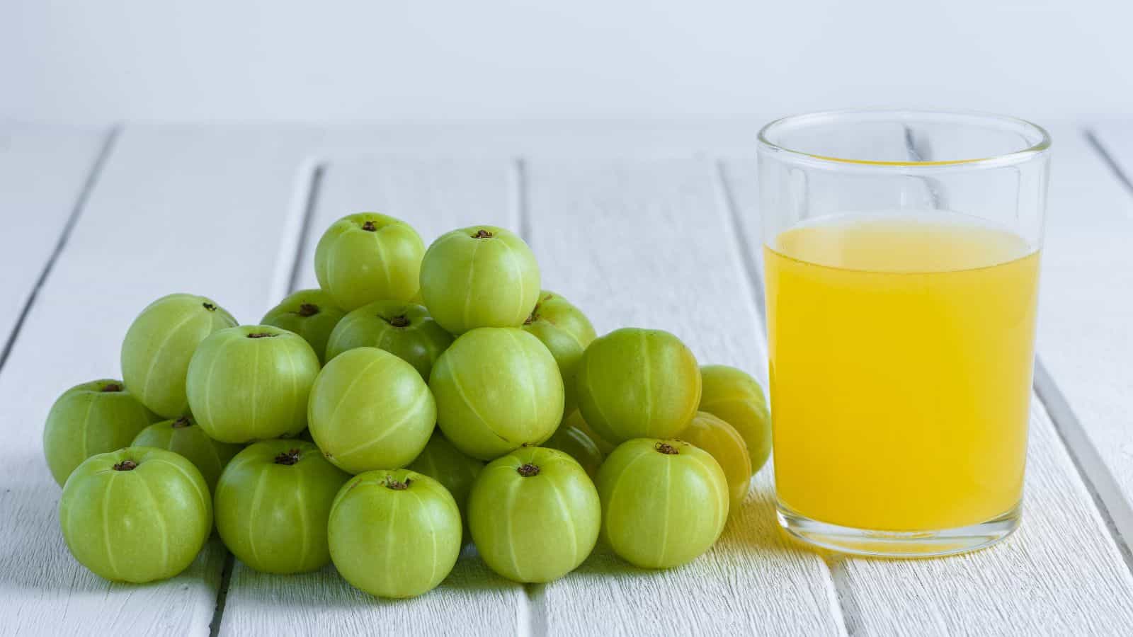 A bunch of fresh green amla fruits is placed next to a glass filled with yellow amla juice on a white wooden surface.