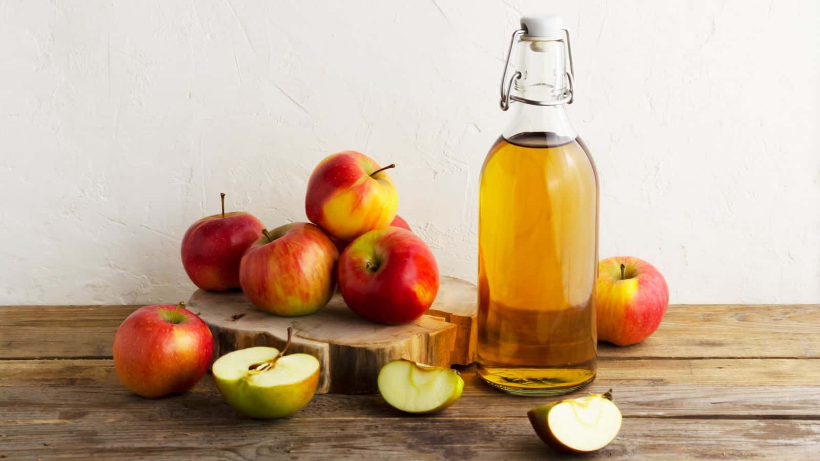 A bottle of apple cider vinegar sits on a wooden surface alongside several red and yellow apples. Some apples are whole, while others are sliced. The background is a plain white wall.