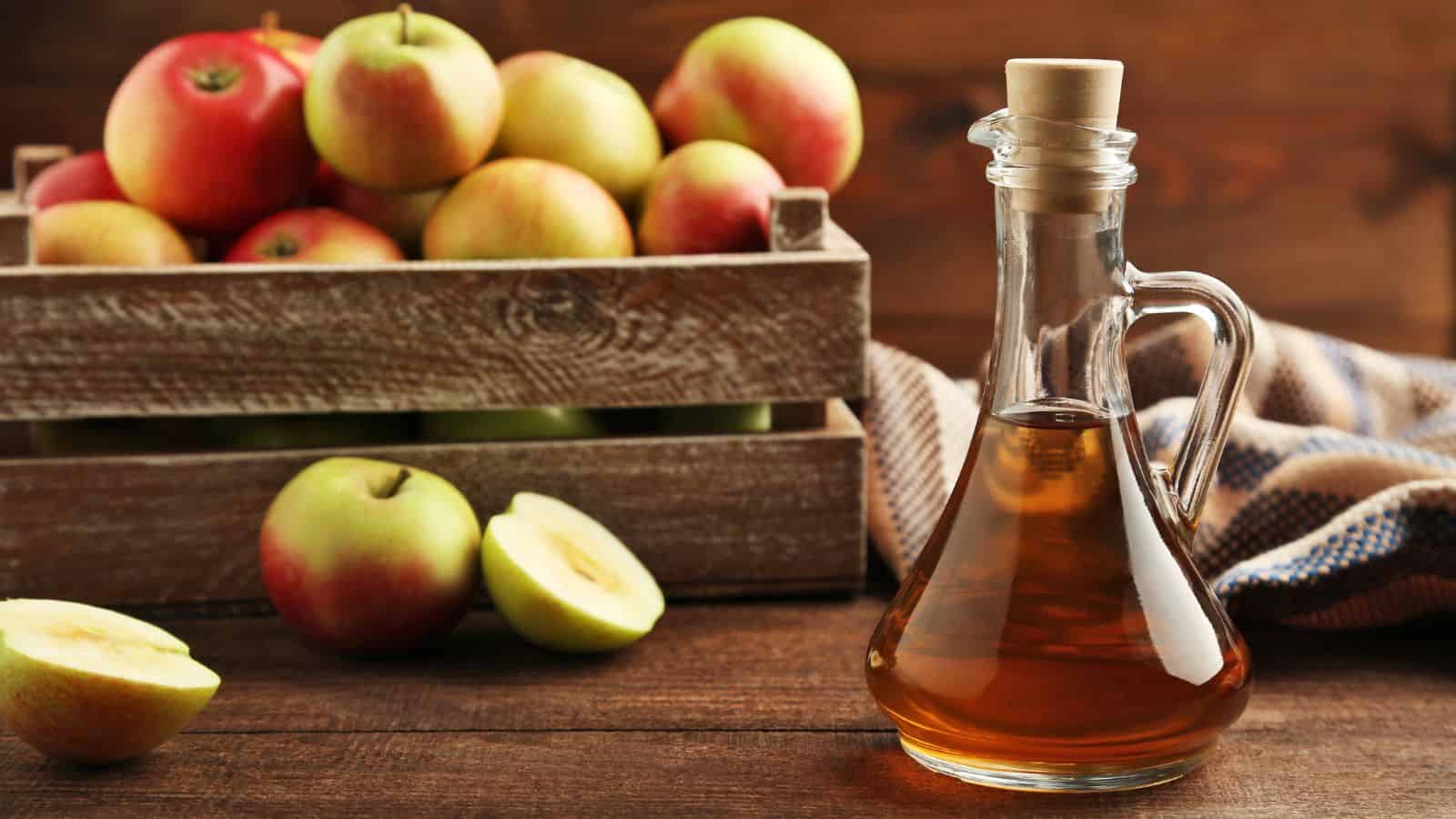 A glass bottle of apple cider vinegar with a cork stopper beside sliced apples on a wooden surface. A rustic wooden crate filled with whole apples is in the background, and a cloth is partially visible to the right.