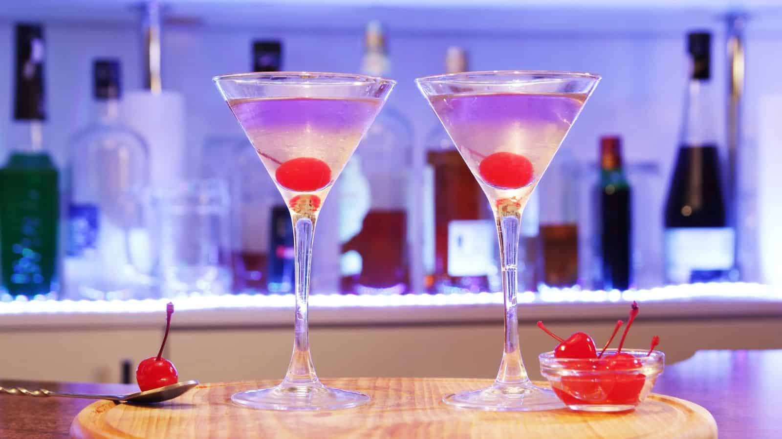 Two martini glasses filled with a clear cocktail and a cherry garnish sit on a wooden surface. Behind them are various liquor bottles on a backlit shelf, creating a colorful bar background. A small bowl of cherries is nearby.