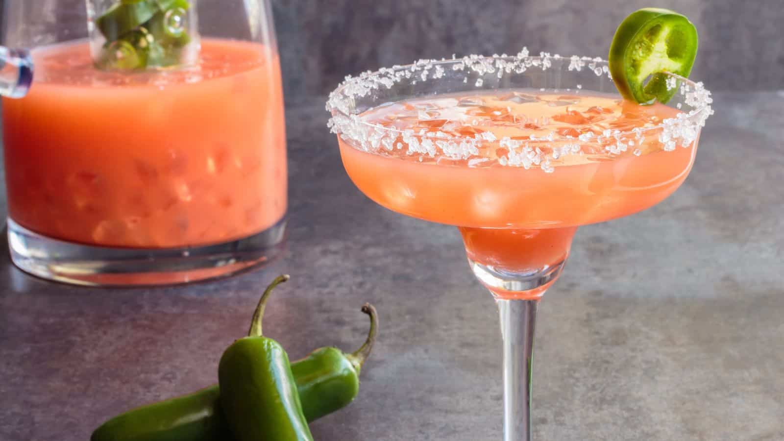 A glass of orange-colored cocktail with a salted rim and a slice of jalapeño is in the foreground. Behind it is a pitcher filled with a similar drink and jalapeño slices. Two whole jalapeños are placed on the surface nearby.