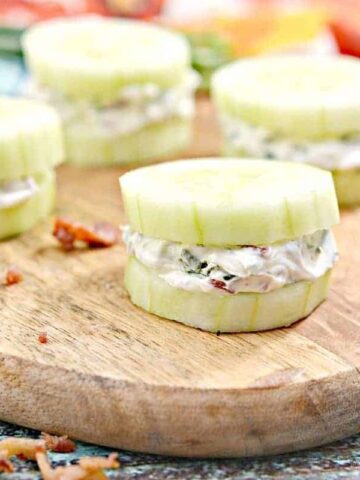 Cucumber sandwich halves on a wooden board, filled with cream cheese and herbs. Bacon bits are scattered around. Bright and colorful ingredients are visible in the background.