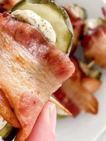 A hand holds an appetizer made with bacon wrapped around a pickle slice and cream cheese, secured with a toothpick. More of these appetizers are visible on a plate in the background, placed on a marble surface.