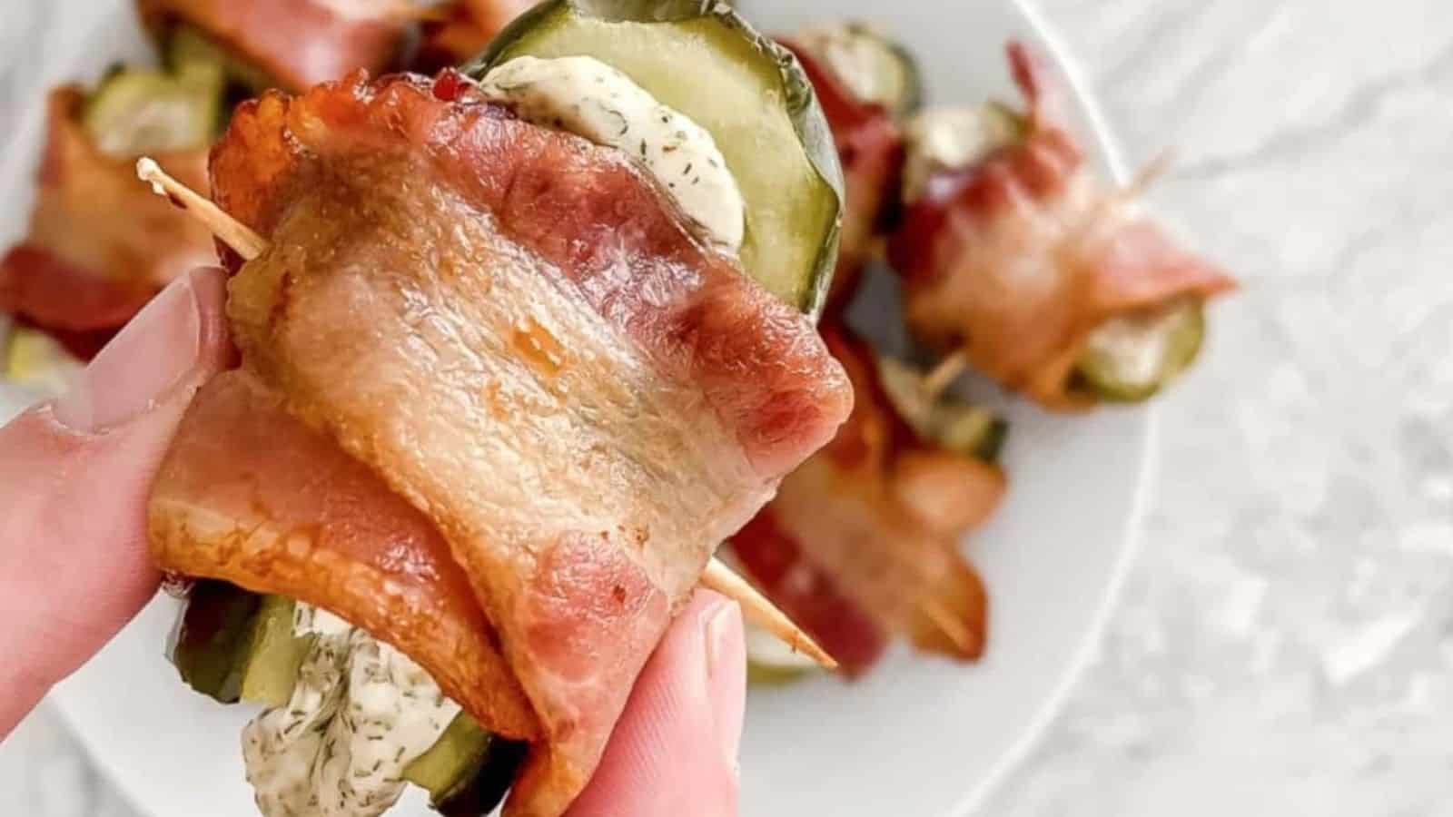 A hand holds an appetizer made with bacon wrapped around a pickle slice and cream cheese, secured with a toothpick. More of these appetizers are visible on a plate in the background, placed on a marble surface.