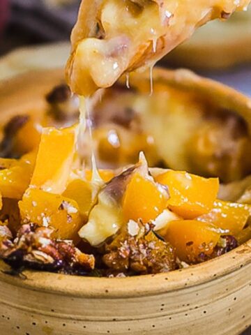 A wooden bowl filled with a cheesy, baked dish containing diced peaches and chopped nuts. A spoon lifts a portion with melted cheese stretching upwards. Slices of bread are visible in the background.