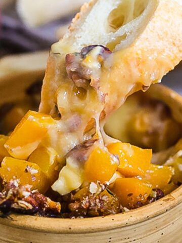 A hand dips a slice of bread into a bowl filled with melted cheese and chopped peaches, creating the perfect appetizer. The dish is topped with crushed pecans. Additional bread slices and pecans are scattered on the table, with a ripe peach visible in the background.