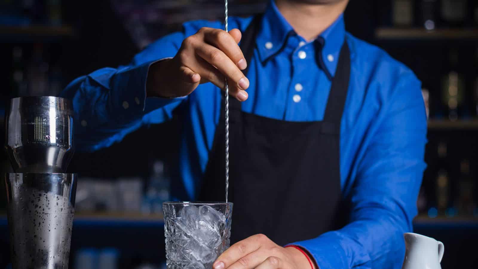 A person in a blue shirt and black apron stirs ice in a glass with a long metal spoon. A cocktail shaker is visible to the left, and a white pitcher is partially visible on the right. The background is dimly lit, suggesting a bar setting.