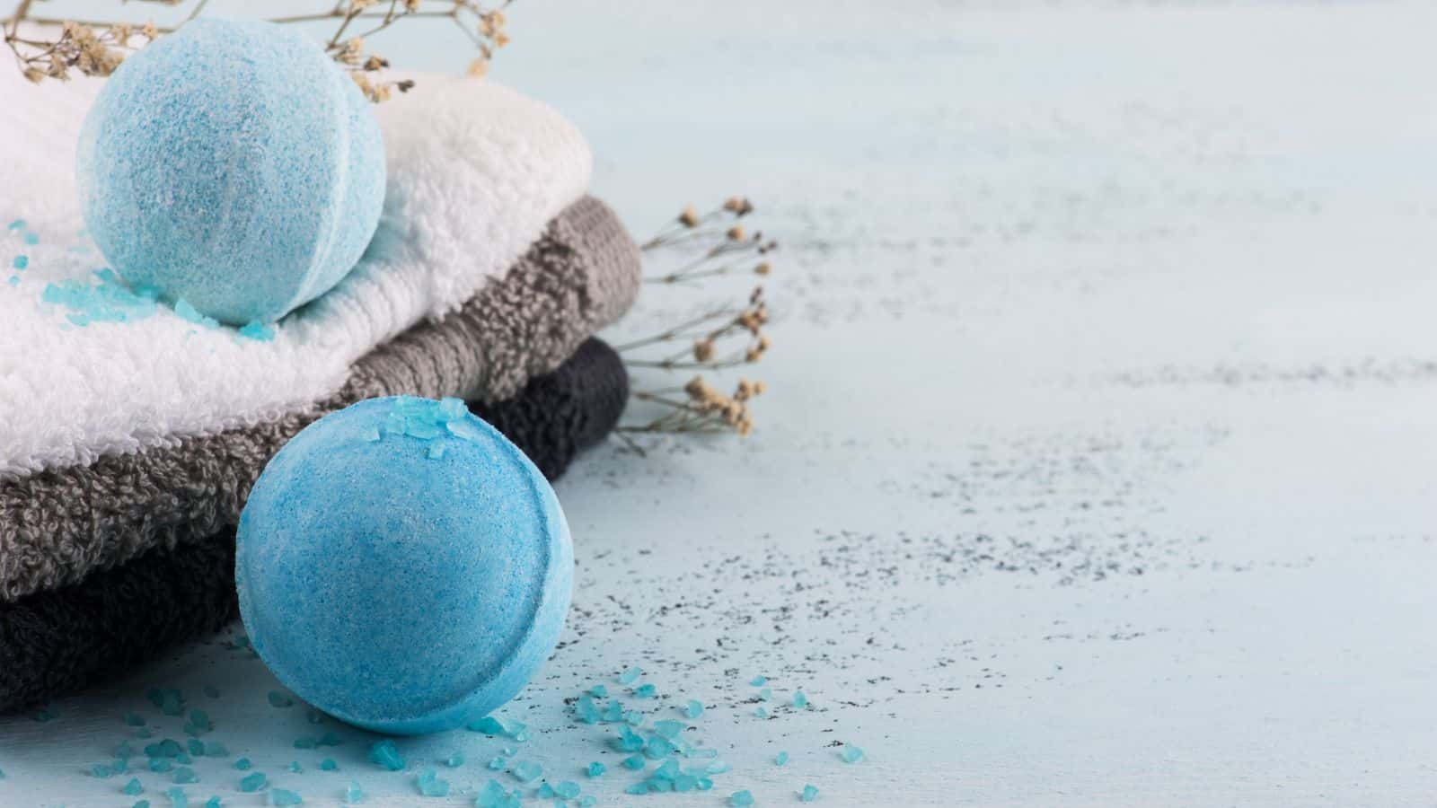 A blue bath bomb on a wooden surface next to a stack of folded towels in white, gray, and black. Blue bath salt crystals are scattered around. Dried flowers are placed nearby for decoration.