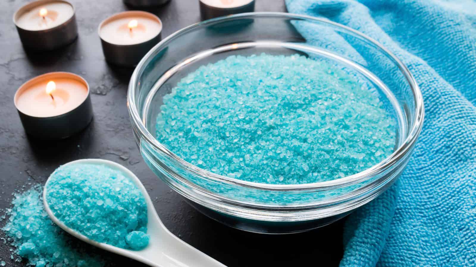 A glass bowl filled with blue bath salts is on a dark surface. A white spoon beside it holds more bath salts. Several small lit candles and a blue towel are in the background.