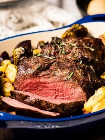 A blue dish filled with sliced roast beef, surrounded by roasted potatoes and vegetables. The beef is cooked medium rare, with a pink center. The dish is placed on a wooden cutting board, and bread rolls are visible in the background.