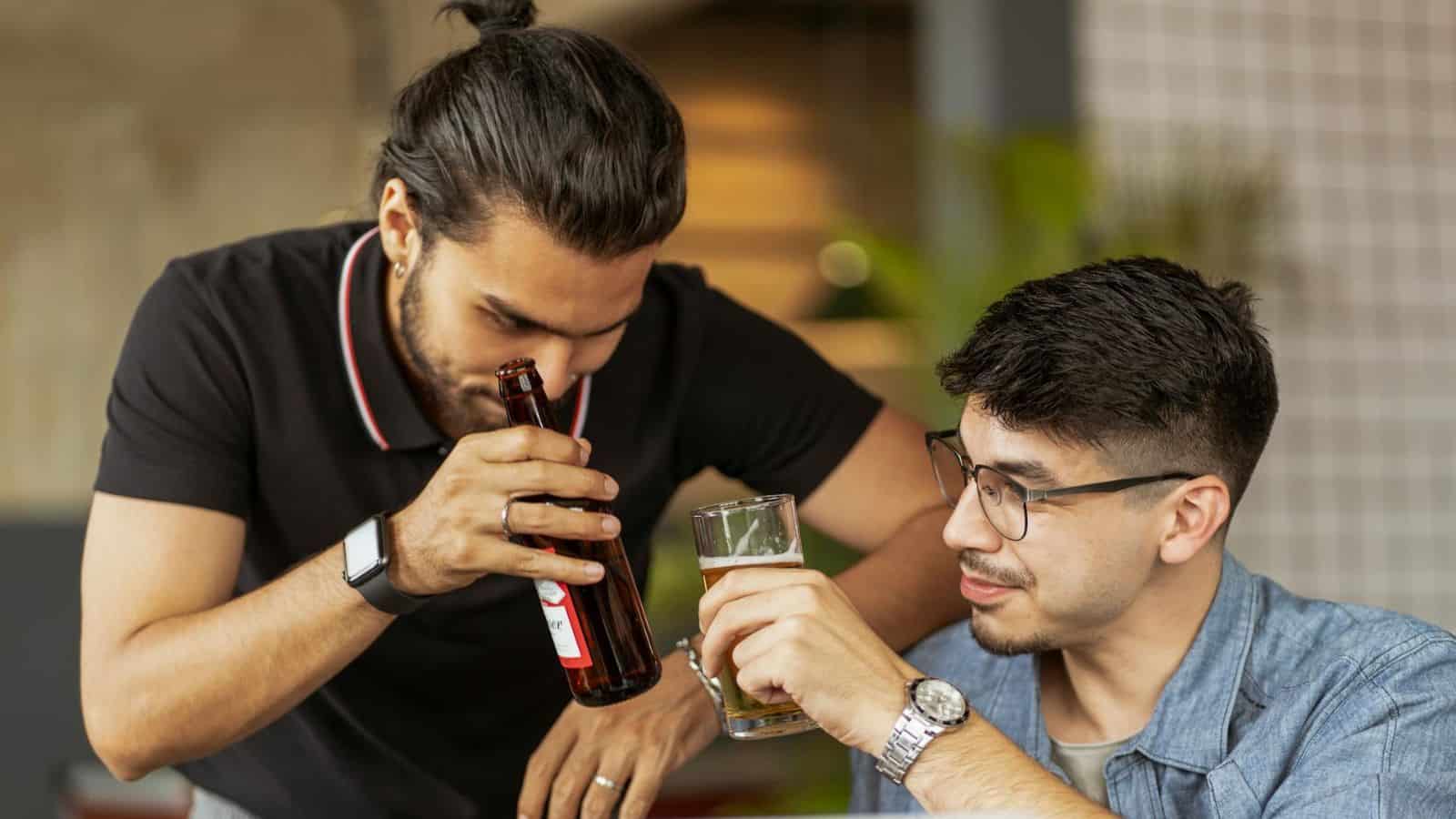 Two men are indoors, one with a bun hairstyle sniffing a beer bottle, and the other wearing glasses holding a glass of beer. Both appear relaxed, engaging in casual conversation at a table.