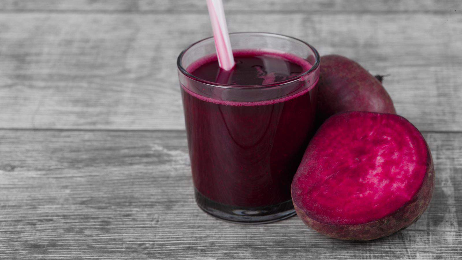 A glass of dark red beet juice with a straw on a wooden surface next to a whole beet and a sliced beet.