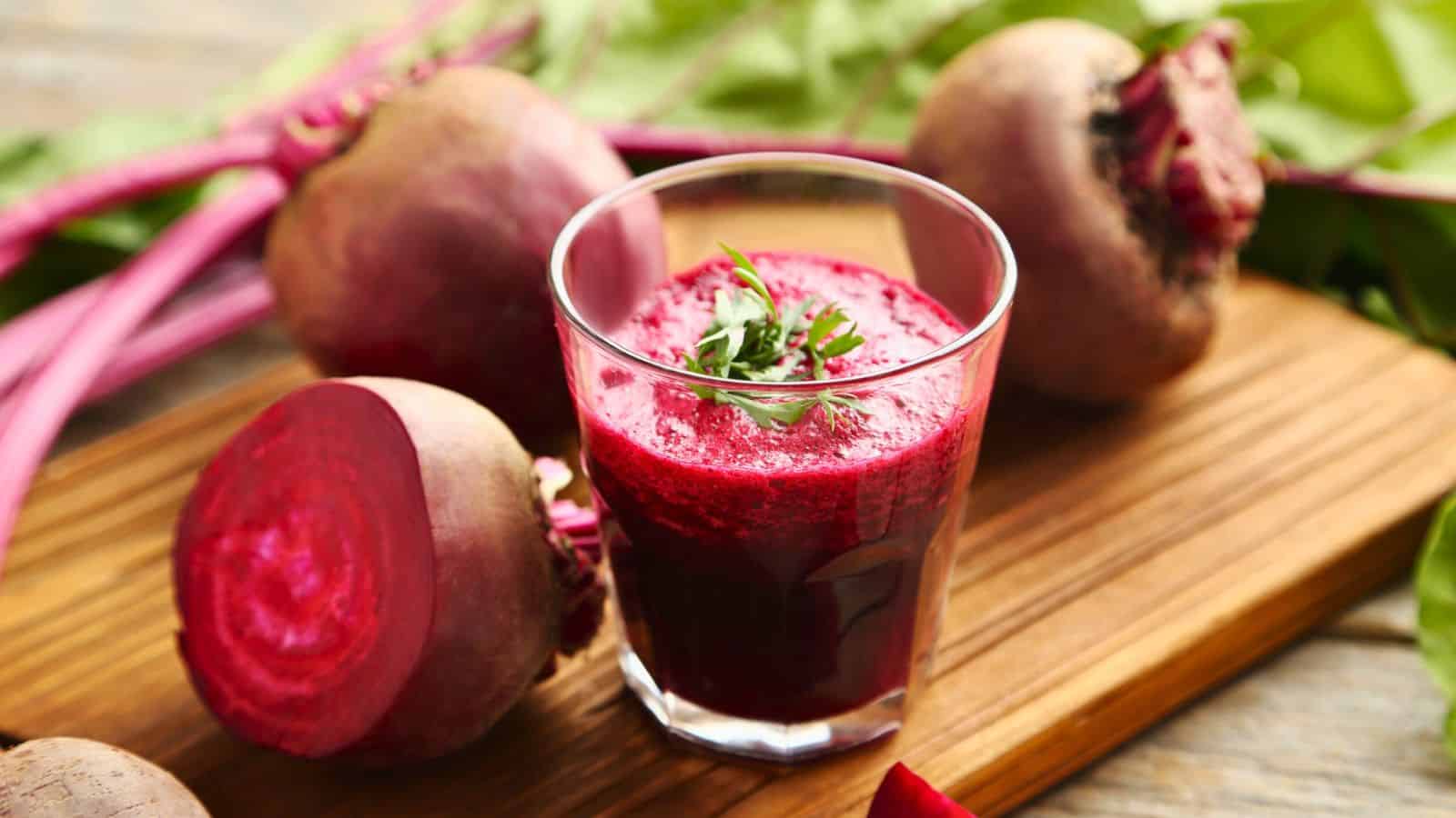 A glass of beetroot juice is placed on a wooden board. Fresh, whole beetroots, one of which is sliced in half, surround the glass. Some green leaves and beet stems are visible in the background.