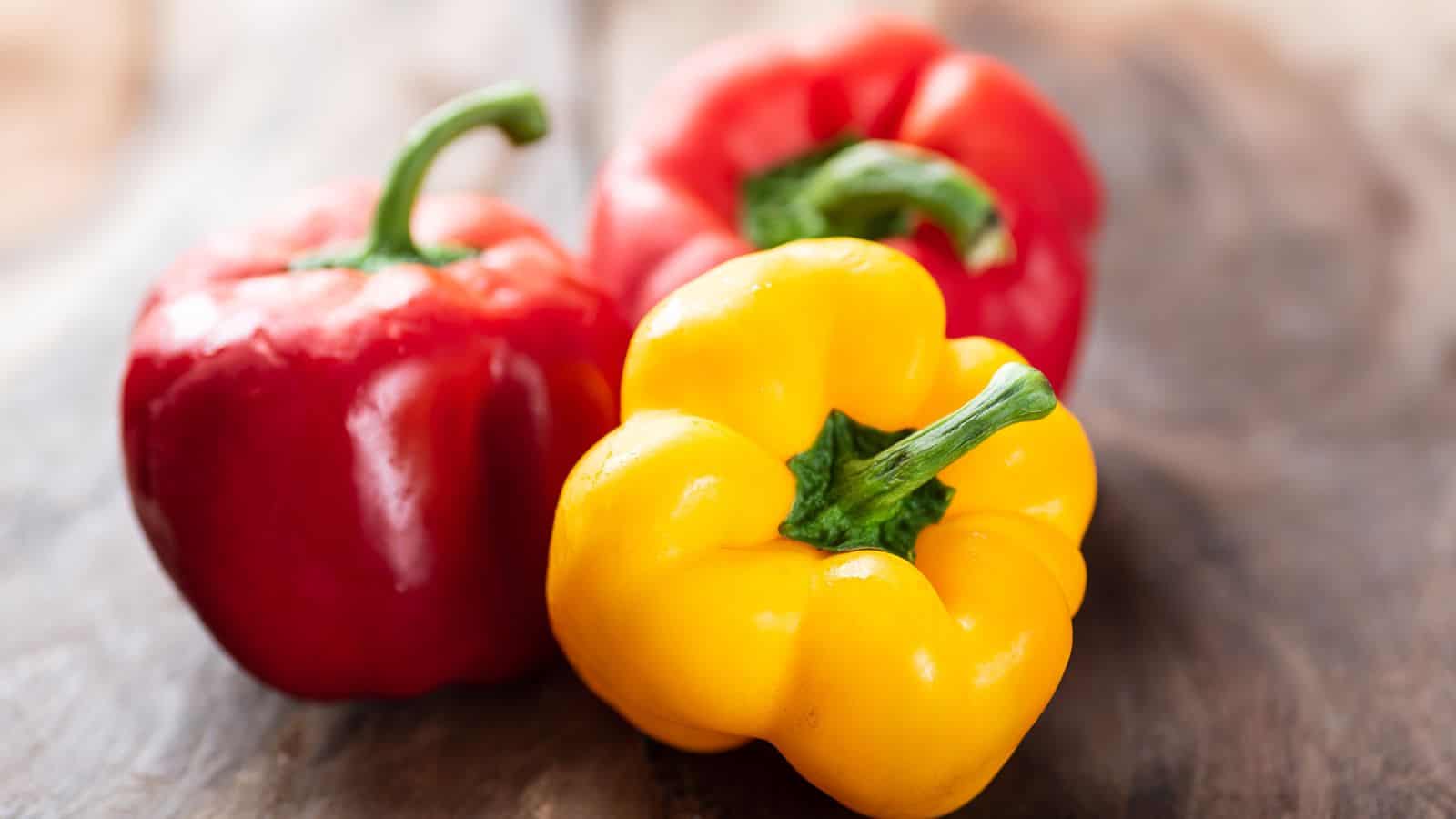 Three bell peppers are shown on a wooden surface. There are two red peppers and one yellow pepper, each with a green stem. The focus is on the front yellow pepper, with the reds slightly blurred in the background.