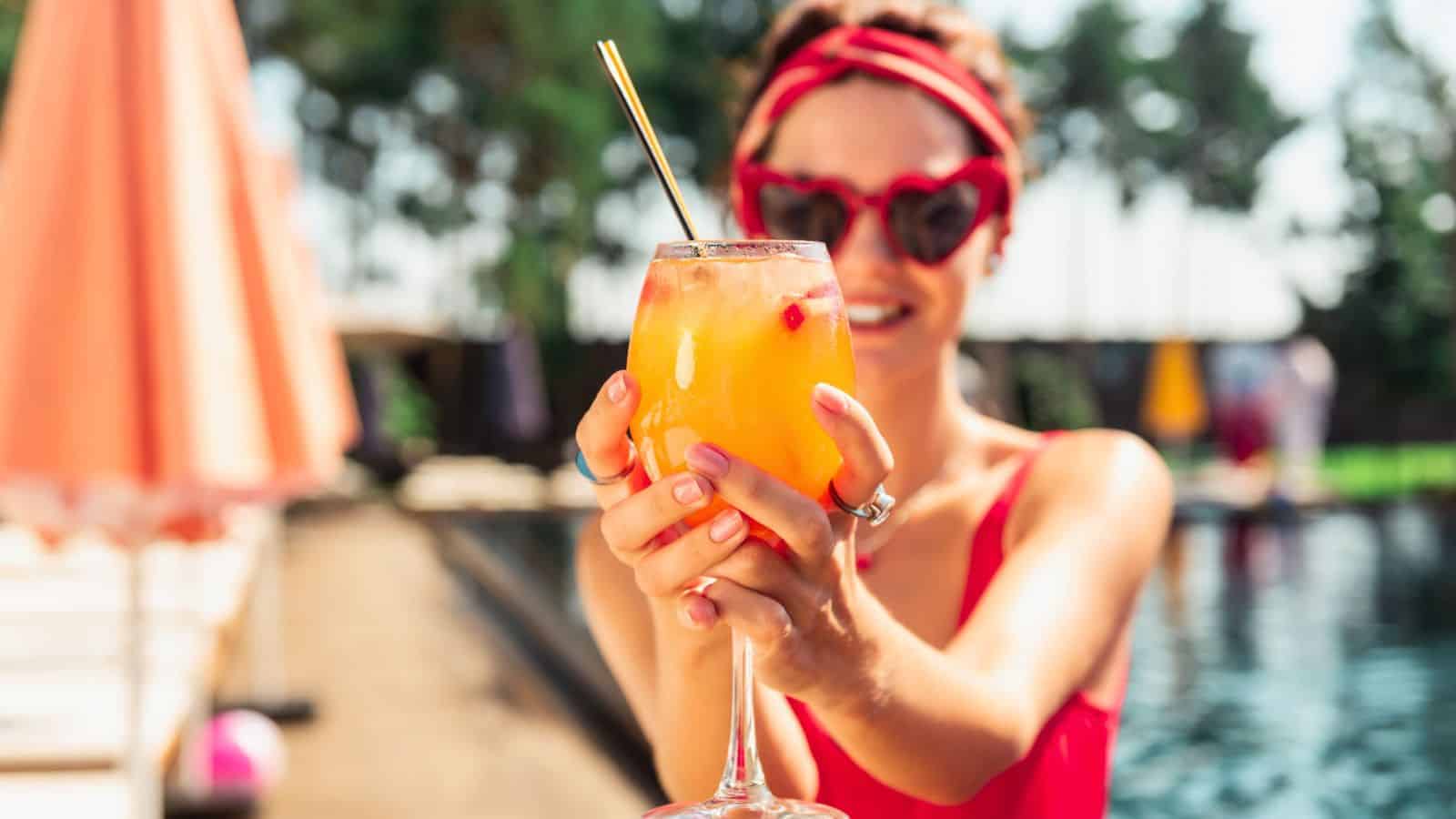 A person wearing a red swimsuit and heart-shaped sunglasses is holding a tropical drink with a straw. They are standing by a poolside, with umbrellas and trees in the background.