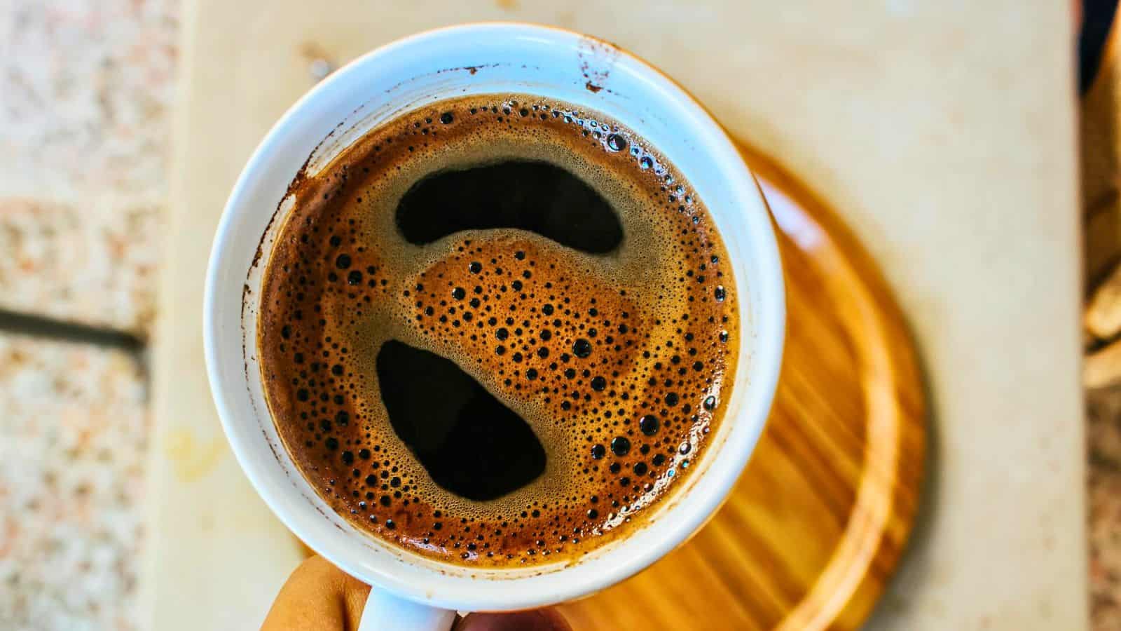 A close-up of a cup of black coffee with a frothy surface, held above a wooden coaster on a tabletop. The coffee appears freshly brewed with visible bubbles on the surface.