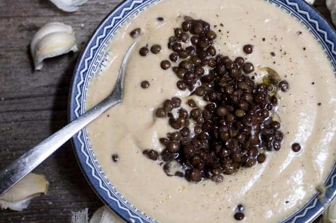 A bowl of creamy soup topped with a generous amount of black lentils, garnished with a drizzle of olive oil and sprinkled with black pepper. A metal spoon rests in the bowl. Garlic cloves are scattered on the wooden surface beside it.