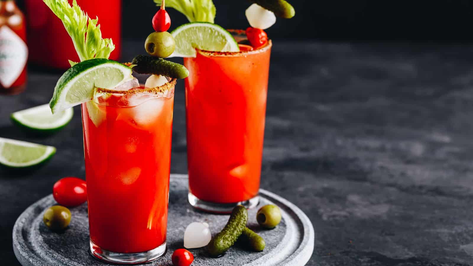 Two tall glasses of Bloody Mary cocktails on a stone tray. Each glass is garnished with celery, lime wedges, green olives, pickles, and cherry tomatoes. A dark background with extra lime wedges and a bottle of hot sauce is visible.