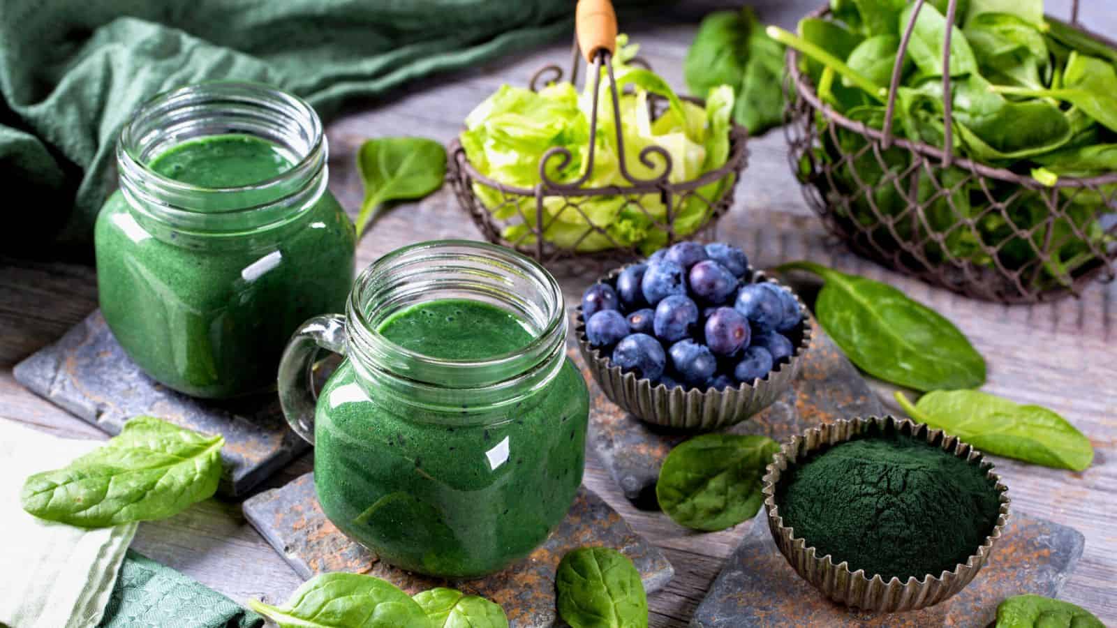 Two jars of green smoothie are on a table surrounded by fresh spinach leaves. There's a small bowl of blueberries, a small dish with green powder, a basket of leafy greens, and a fabric napkin. The setting has a rustic appearance.