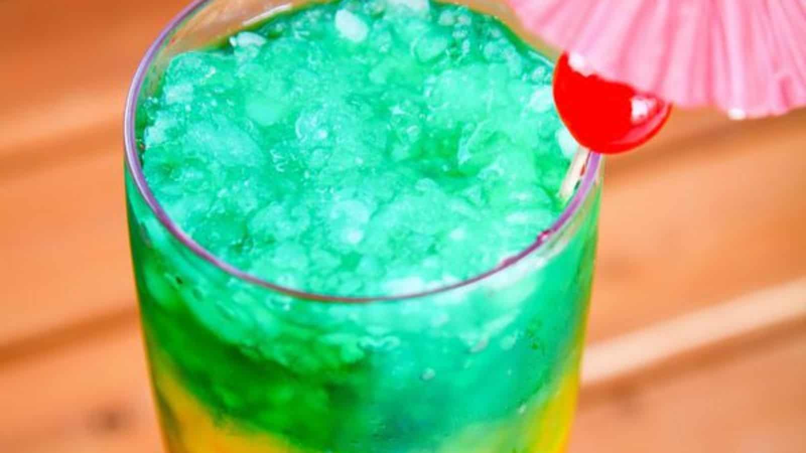 A red yellow and green cocktail garnished with a cherry and cocktail umbrella.