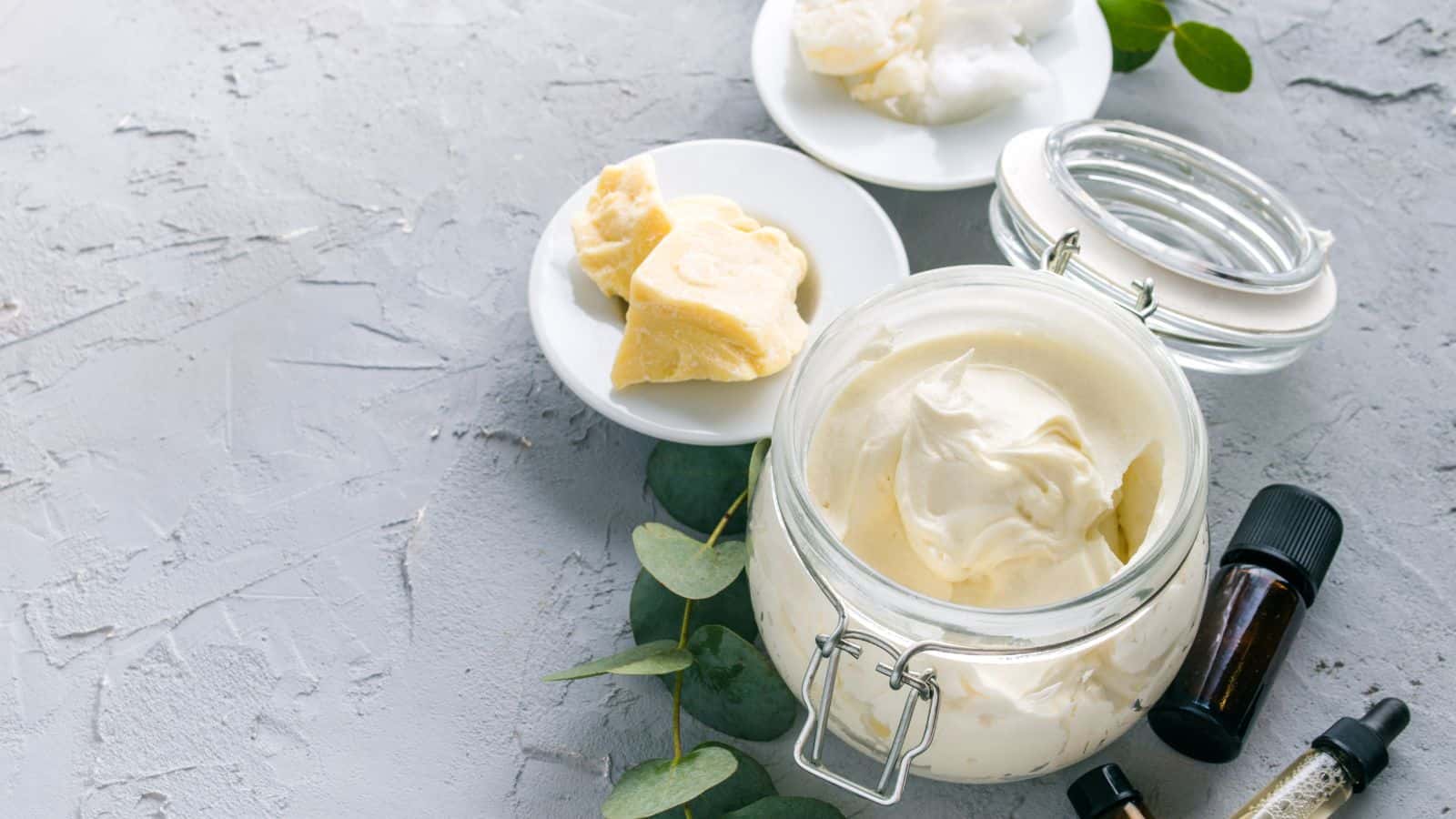 A glass jar filled with a creamy substance is placed on a textured gray surface. Nearby, there are small plates with chunks of solid ingredients, two small dark glass bottles, and green leaves scattered around.