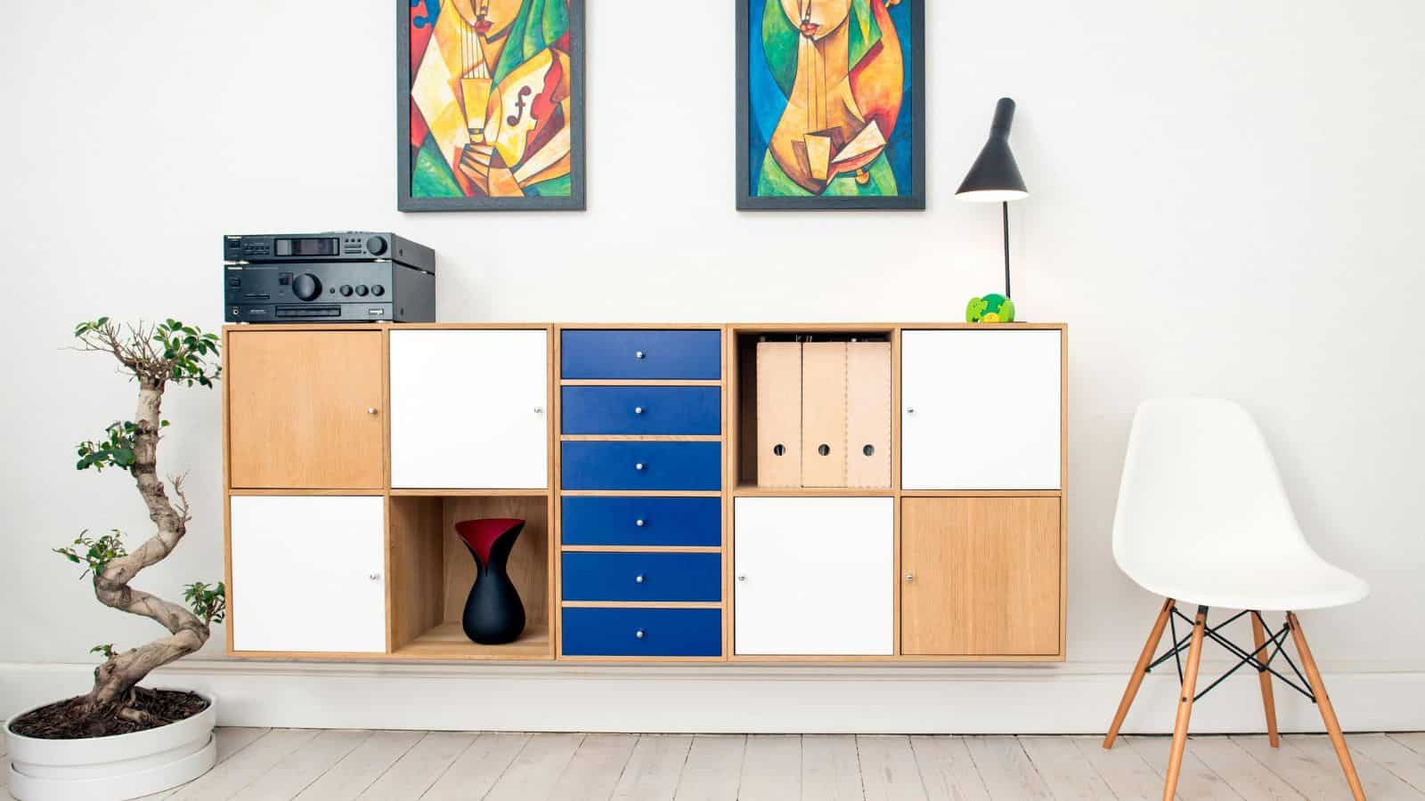 A modern living room features a wooden cabinet with white and blue doors, holding a stereo system and various items. Above, two colorful abstract paintings hang on the wall. A small tree in a pot and a white chair complete the scene.