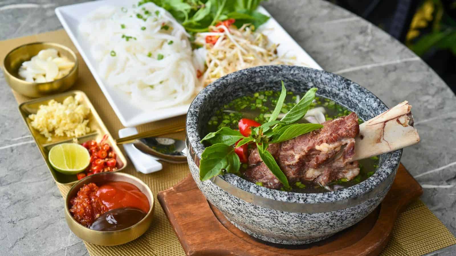 A bowl of soup with beef bones and herbs is placed on a wooden base. A plate with fresh noodles and greens is in the background, alongside small dishes containing various condiments like ginger, lime, and chili sauce.