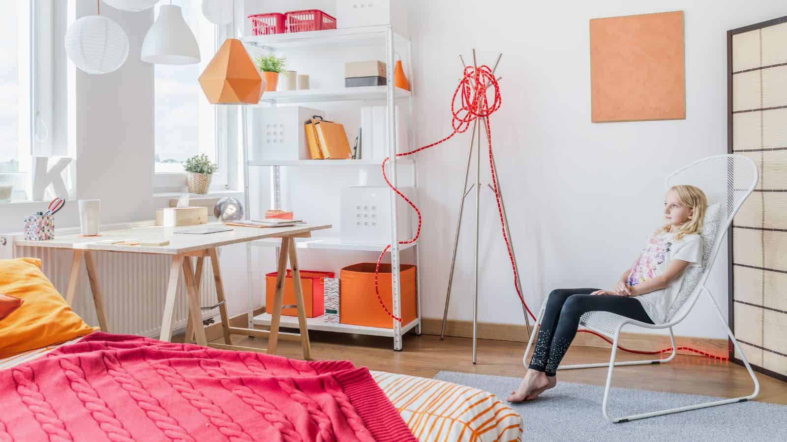 A child sits on a white chair in a bright room with modern decor. The room features a bed with colorful bedding, a shelving unit with decor items, a desk by the window, and decorative orange and red elements. A window allows natural light in.