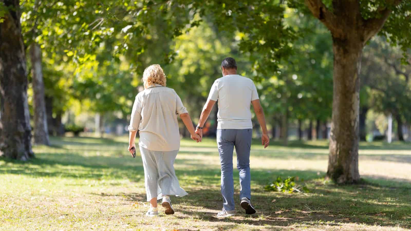 A couple holding hands walks away down a tree-lined path in a park. The woman on the left has shoulder-length blonde hair and wears a light outfit. The man on the right has short hair and wears a white shirt and gray pants. The sun filters through the trees.