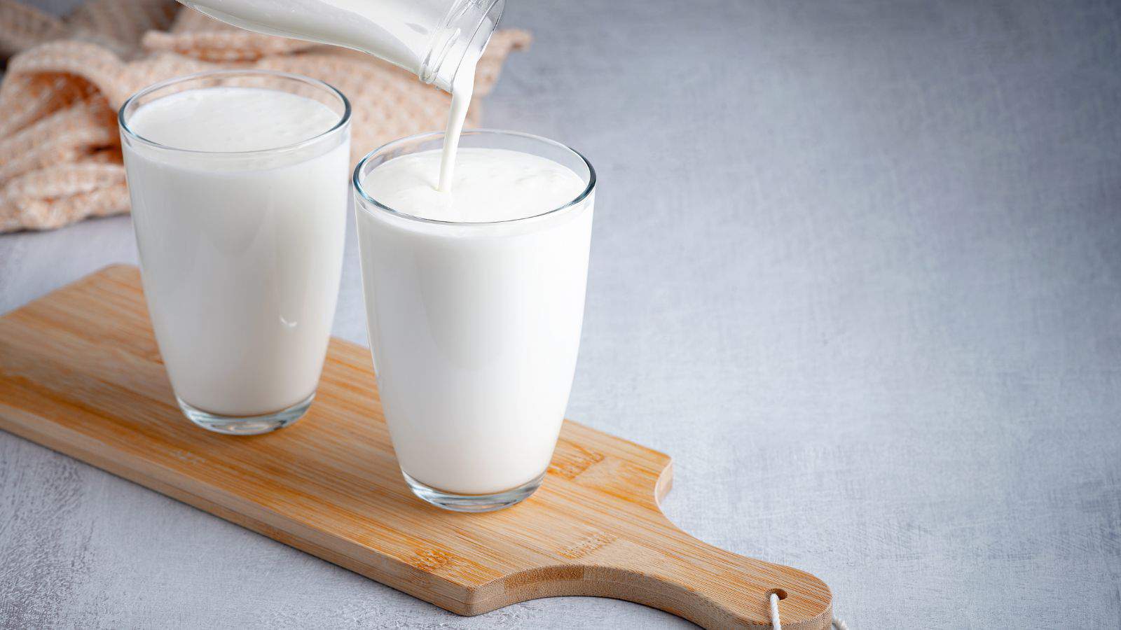 Two glasses filled with milk are placed on a wooden cutting board. A bottle is pouring milk into one of the glasses. A beige cloth is in the background on a gray surface.