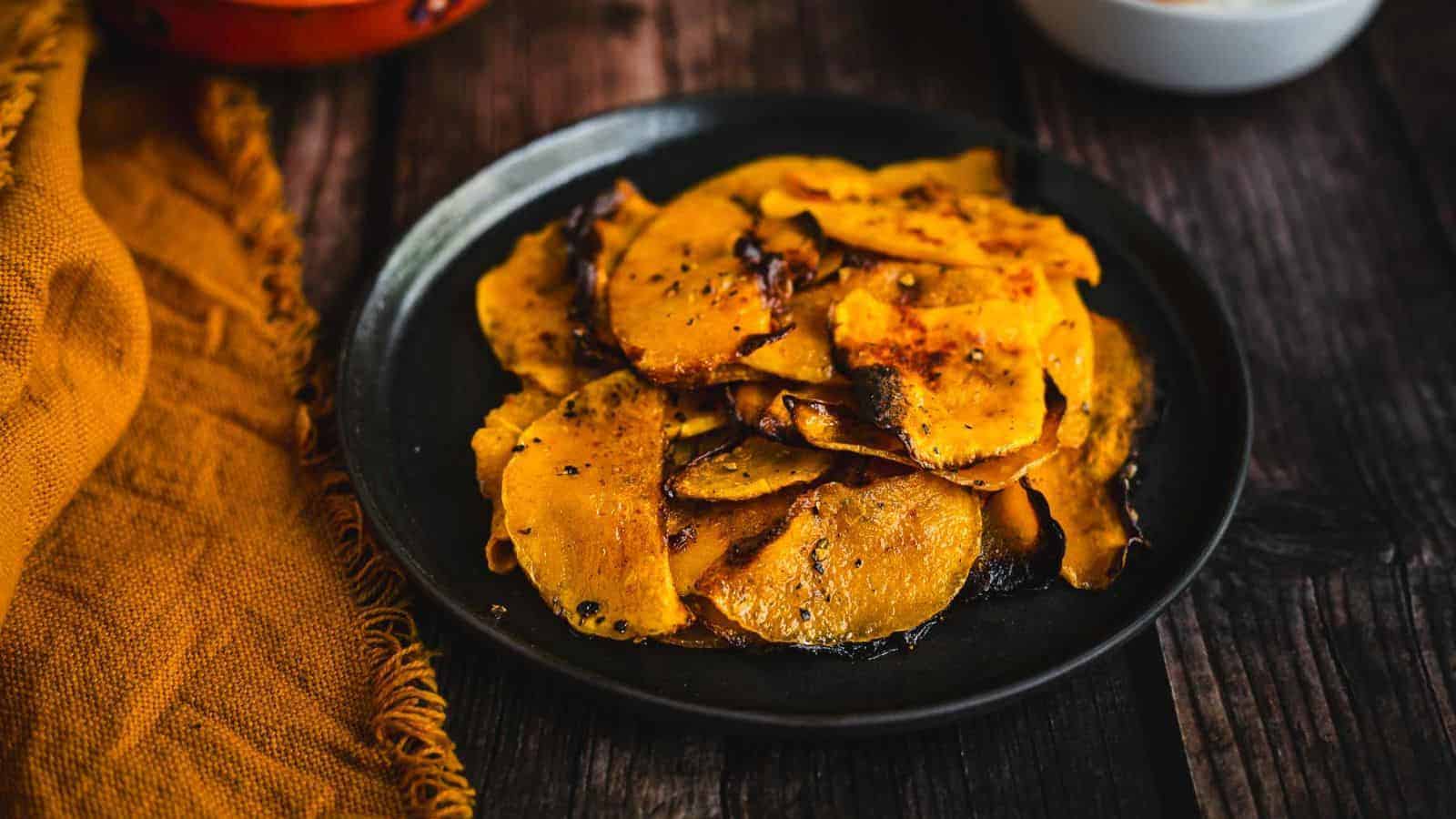 A plate of roasted butternut squash slices, seasoned and browned, is set on a wooden table. A mustard yellow cloth is partially visible on the left side of the image.